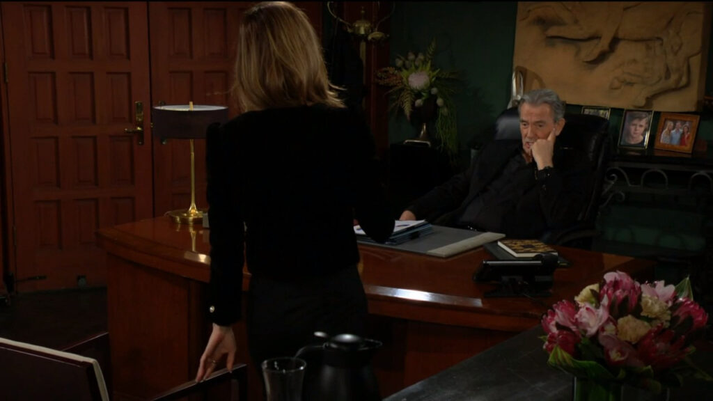 Victor sits behind the desk while he talks to Victoria