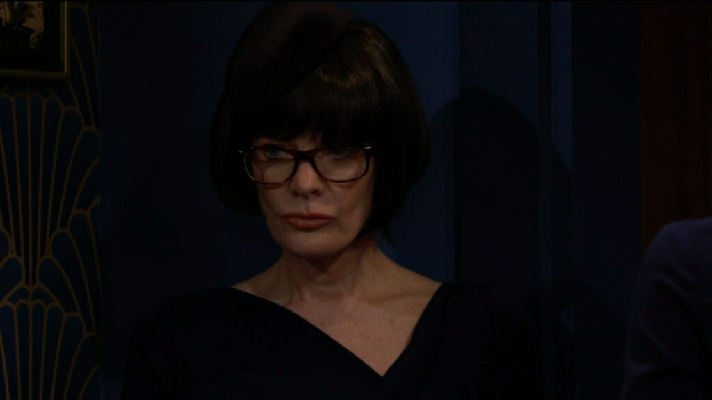Phyllis, disguised in a wig and glasses, stands in the back of the room observing the memorial service