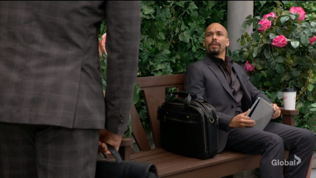 Devon sits on a park bench with a tablet in hand and a briefcase beside him. He looks up at Nate as they talk.