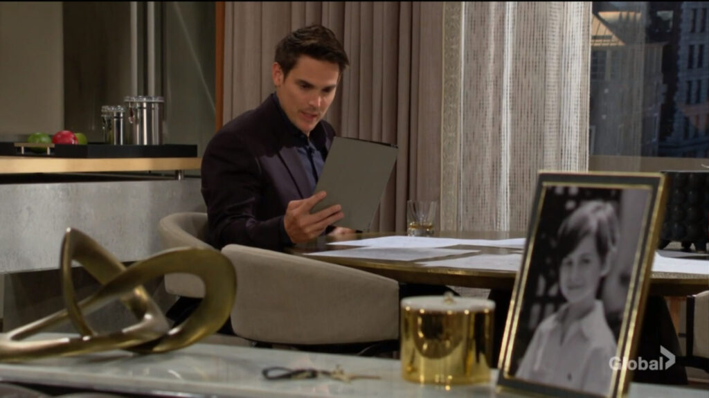 Adam sits and looks at his tablet. There's a picture of Connor on the sideboard.